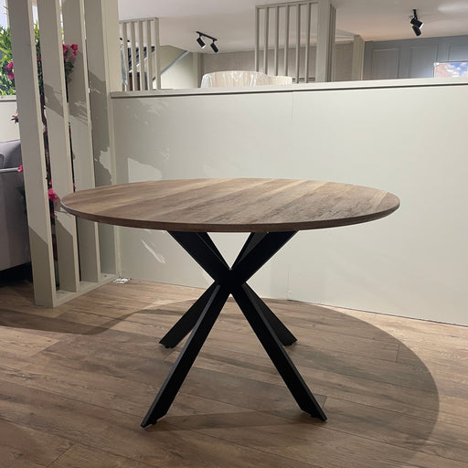 The Elson Round Dining Table, featuring a light wood top and sleek black X legs, is set on a wooden floor in a modern, well-lit room with neutral walls and vertical decorative slats.