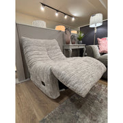 The Ryder Occasional Chair, featuring a gray textured finish, 360-degree swivel, and curved design, is displayed in a showroom with wooden floors, vases, a gray sofa, and a standing lamp in the background.
