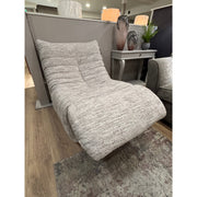 The Ryder Occasional Chair, a modern light gray upholstered lounge with a curved reclined shape, sits on a wooden floor beside a gray side table with decorative vases and a plant. A gray sofa and area rug are partially visible nearby.
