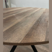 A close-up of the Elson Round Dining Table showcases its visible grain patterns and smooth surface, while modern black X legs stand out. The blurred background features a white wall and part of a shelf.