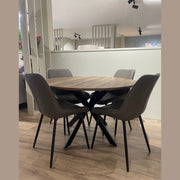 The Elson Round Dining Table with a black metal base and modern style is paired with four gray upholstered chairs with black legs, set on a light wooden floor in a contemporary interior.