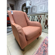 The Brontë Occasional Chair, featuring a modern salmon hue, wooden legs, and timeless design, is showcased in a showroom with tiled floors, decorative wall panels, and mirrored cabinets in the background.