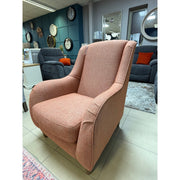 The Brontë Occasional Chair, upholstered in salmon-pink with cushioned seat and back and subtly curved arms for a timeless look, is displayed on a tiled showroom floor among other furniture, wall clocks, and a narrow arm sofa.