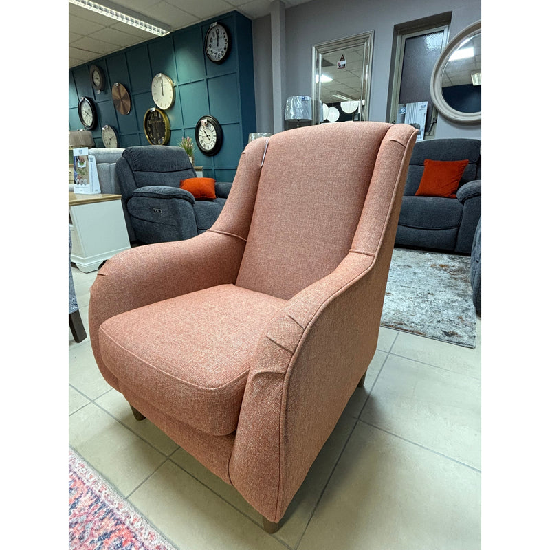 The Brontë Occasional Chair, upholstered in salmon-pink with cushioned seat and back and subtly curved arms for a timeless look, is displayed on a tiled showroom floor among other furniture, wall clocks, and a narrow arm sofa.