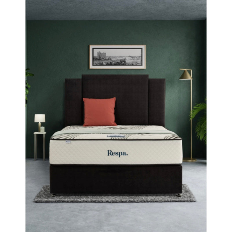 A modern bedroom with a dark upholstered bed, a cream Respa Jupiter mattress, one orange pillow, two side tables with lamps, a floor lamp, a framed picture, and a plant against a dark green wall.