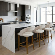 Modern kitchen with a large white marble island, four Willow Counter Stools featuring beige upholstery and wooden legs, dark wood cabinets, stainless steel appliances, and large windows letting in natural light.
