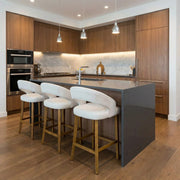 Modern kitchen with wood cabinets, marble backsplash, stainless steel appliances, and a gray island with sink. Four white Willow Counter Stools are lined up at the island beneath three pendant lights.