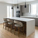 Modern kitchen featuring a large white marble island, three Willow Counter Stools, light wood floors, sleek gray cabinets, built-in ovens, white tile backsplash, and three black pendant lights above the island.