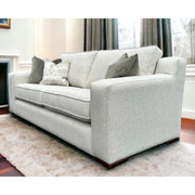 The Alice Range, a light gray suite with modern square arms and four throw pillows, is featured in a bright living room with large windows, dark curtains, and a plush white rug. Made in Ireland.