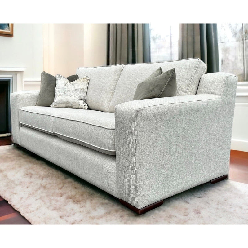 The Alice Range, a light gray suite with modern square arms and four throw pillows, is featured in a bright living room with large windows, dark curtains, and a plush white rug. Made in Ireland.