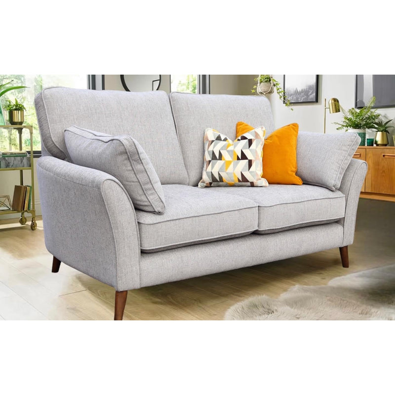 The Retro Sofa Range in light gray with beech frames and wooden legs, accented by patterned and solid yellow pillows, sits in a bright living room featuring plants, wooden furniture, and large windows.