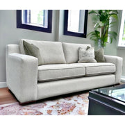 A beige fabric sofa with a modern square-shaped arm, displayed in a room with a patterned rug and windows in the background.