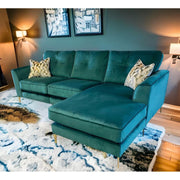 A modern living room showcases the Irish-made Turino Chaise in green aquaclean velvet with gold legs, two patterned throw pillows, abstract wall art, wall-mounted lighting, and a blue and white patterned rug on a wooden floor.