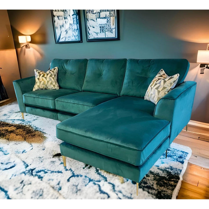 A modern living room showcases the Irish-made Turino Chaise in green aquaclean velvet with gold legs, two patterned throw pillows, abstract wall art, wall-mounted lighting, and a blue and white patterned rug on a wooden floor.