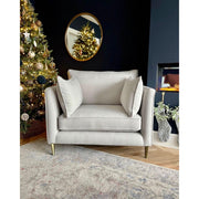 The Hugo armchair in cozy light gray aquaclean fabric with two cushions sits on a patterned rug near a Christmas tree. A round mirror on the dark wall reflects the tree, while a glass vase and plants add decor nearby.