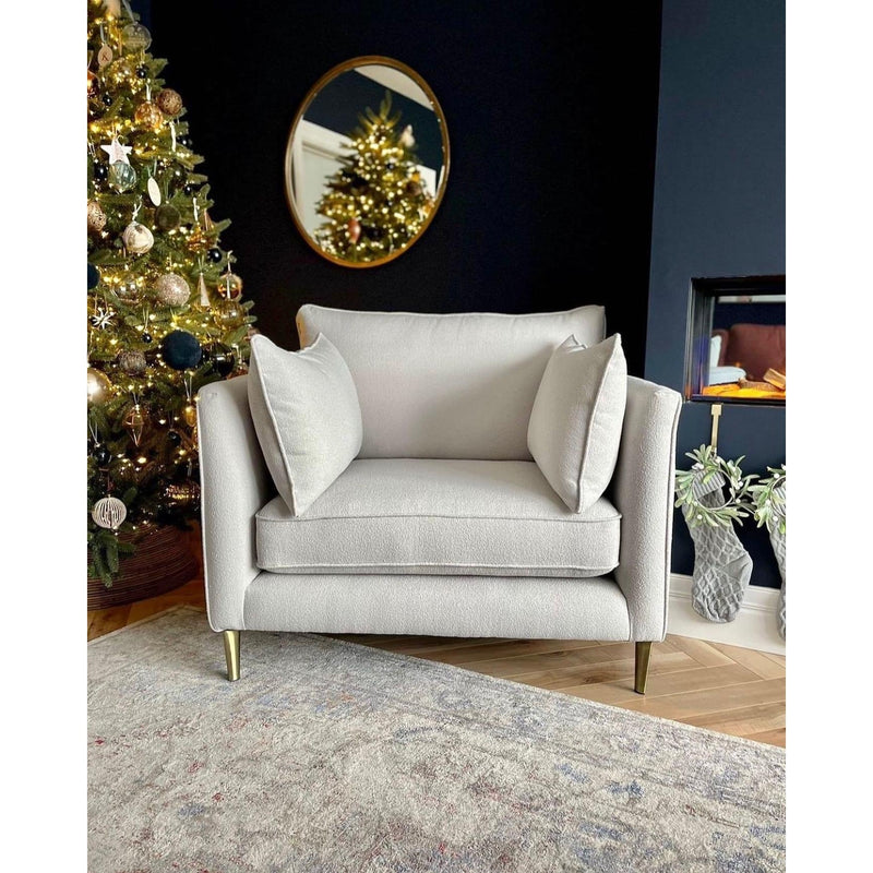 The Hugo armchair in cozy light gray aquaclean fabric with two cushions sits on a patterned rug near a Christmas tree. A round mirror on the dark wall reflects the tree, while a glass vase and plants add decor nearby.