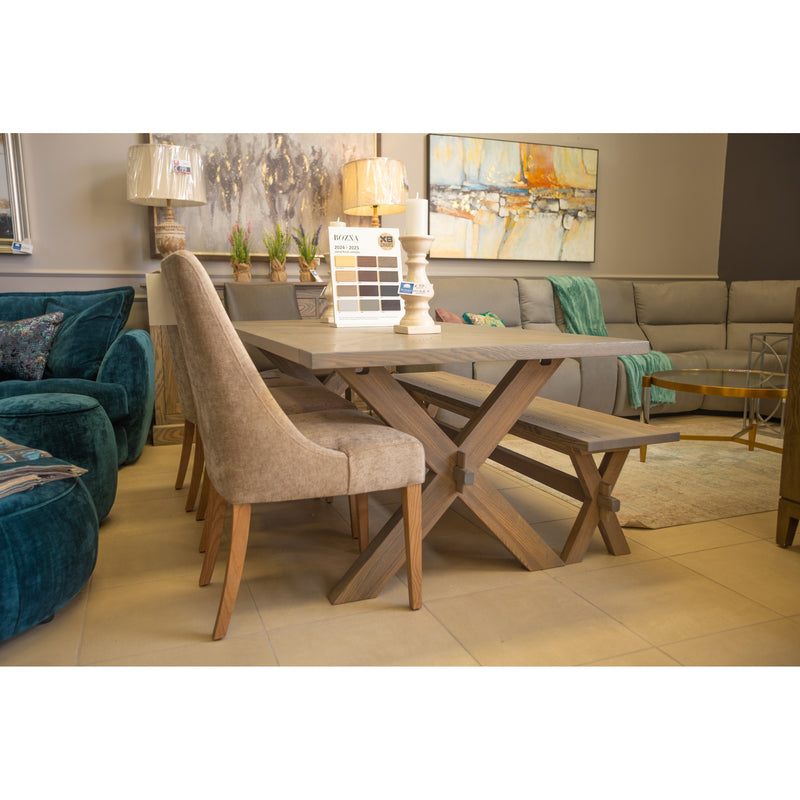 A modern living room showroom featuring the Audrey Dining Table by vendor-unknown, beige upholstered chairs, a bench, gray sectional sofa, colorful abstract paintings, and decorative items on the table.