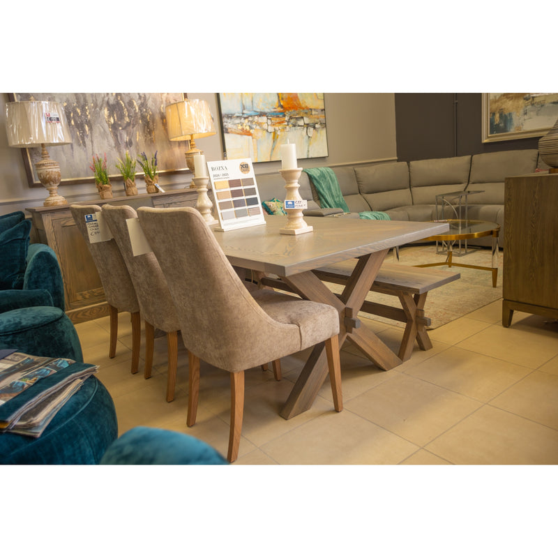 A modern showroom showcases the Audrey Dining Table by vendor-unknown with six beige upholstered chairs, decorative candles, and art pieces. Nearby, a teal armchair and a contemporary sectional sofa complete the space.