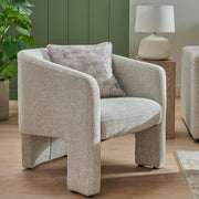 The Humphrey’s Occasional Chair, a modern beige accent with wooden legs and rounded edges, sits on a light wood floor. Its textured chenille cushion is paired with green wall paneling, a potted plant, and a lamp on a wooden side table in the background.