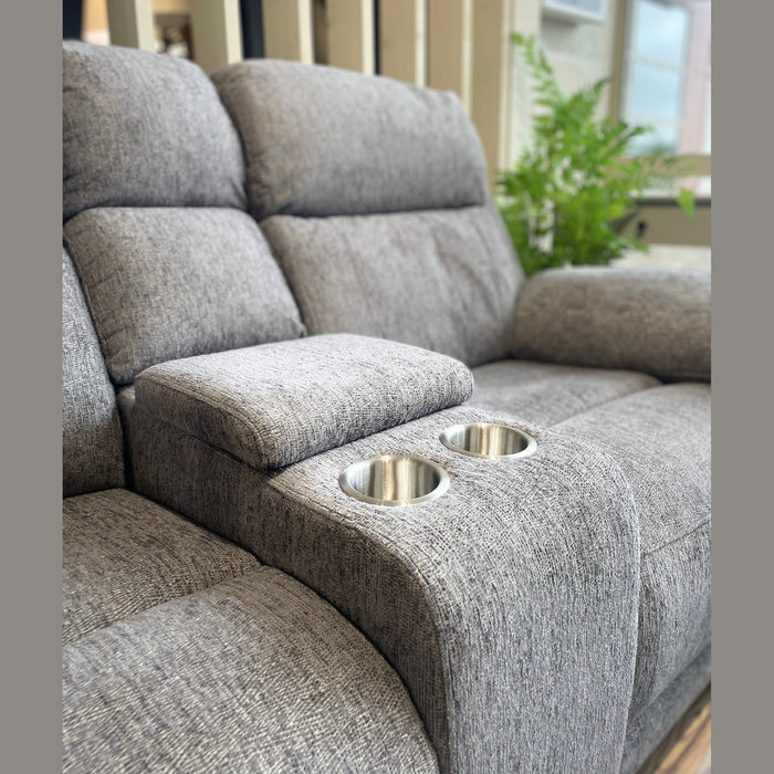 The Elenor Electric Reclining Range is a gray upholstered loveseat with a center console, two cup holders, and padded armrest in luxury fabric, placed indoors by a window with a potted fern in the background.