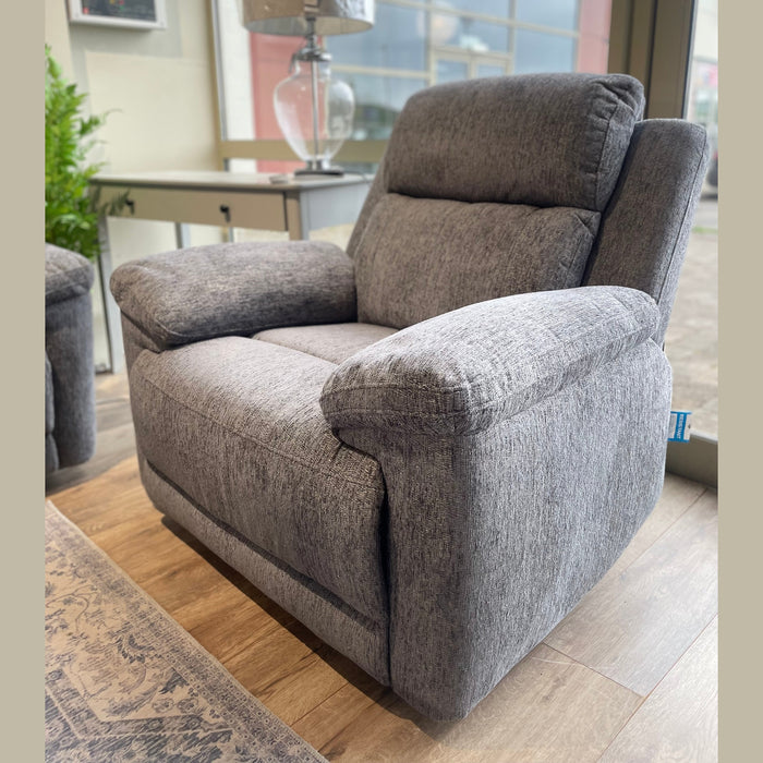 An Elenor Electric Reclining Range armchair in plush gray upholstery sits by a window on a wooden floor, with a clear glass-based lamp, a small table, and a green potted plant in the background.