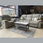 A modern living room showroom features a Reno Recliner Sofa in washable fabric, a matching gray 3-seater with floral cushions, a gray coffee table with drawer, and a patterned rug on tiled floors. Large windows fill the space with natural light.