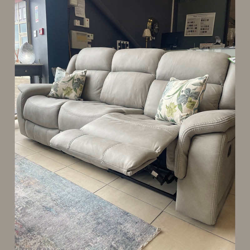 The Reno Recliner Sofa, a modern light gray 3-seater, is shown in a showroom with two floral throw pillows. The middle seat is reclined with the footrest extended. Its washable fabric pairs well with stylish rugs and the décor.
