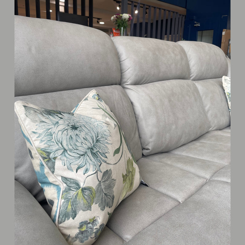 A close-up of the Reno Recliner Sofa, a light gray upholstered 3-seater with a blue and green floral cushion. The background features a blurred modern interior with flowers on a table. The sofa's washable fabric allows for easy care.