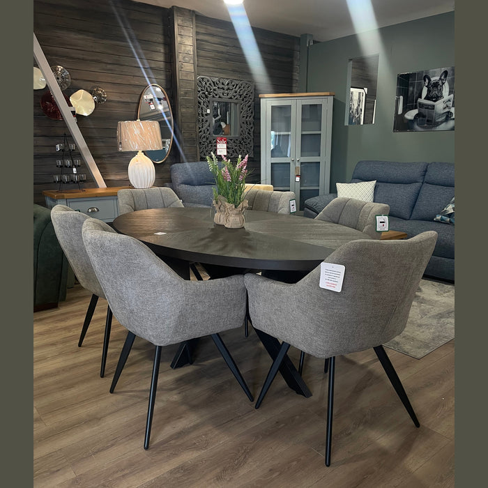 A modern dining room showcases the Samara Oval Table, scratch-proof and paired with six gray upholstered chairs on light wood flooring. A decorative plant sits atop the table, with sofas, cabinets, and decor in the background.