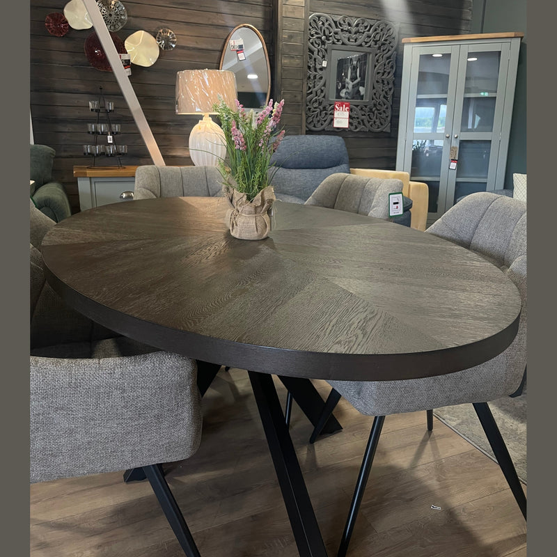 A modern showroom features the Samara Oval Table with a potted plant centerpiece, surrounded by six gray cushioned chairs. Decorative wall art, a lamp, and a mirror complete the elegant setting.