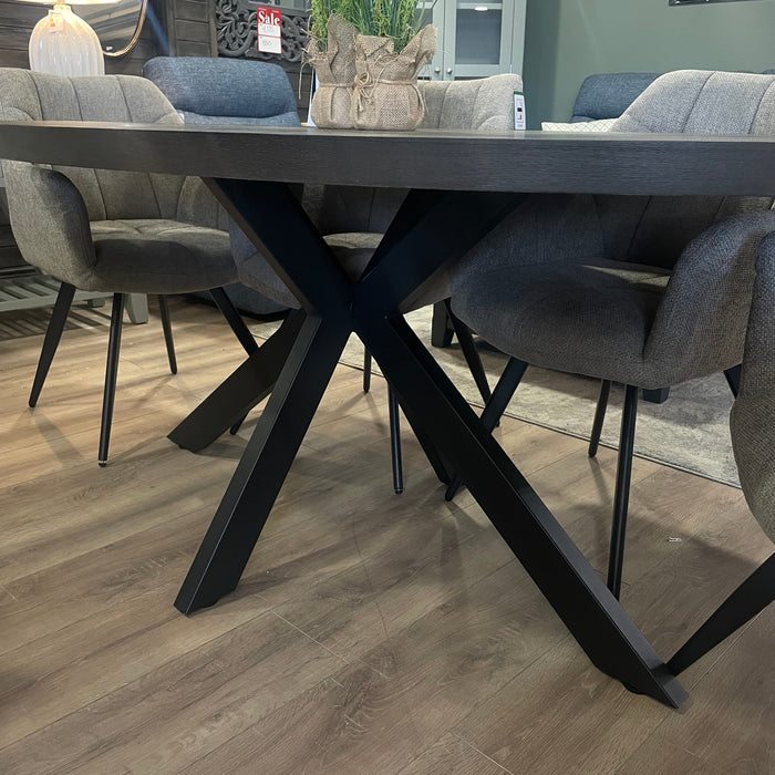 A modern dining room with a Samara Oval Table featuring black metal crossed legs, surrounded by gray upholstered chairs on a light wood floor. A vase with flowers decorates this elegant dining table.