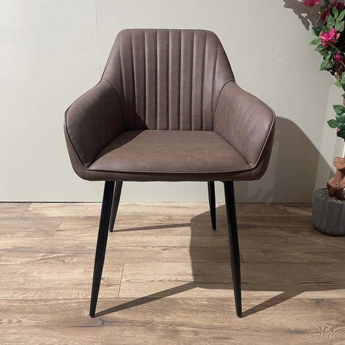 The Enzo Dining Chair features brown leatherette upholstery, vertical stitching, and cushioned seats. It is placed on a wooden floor beside a potted plant with pink flowers and set against a light-colored wall.