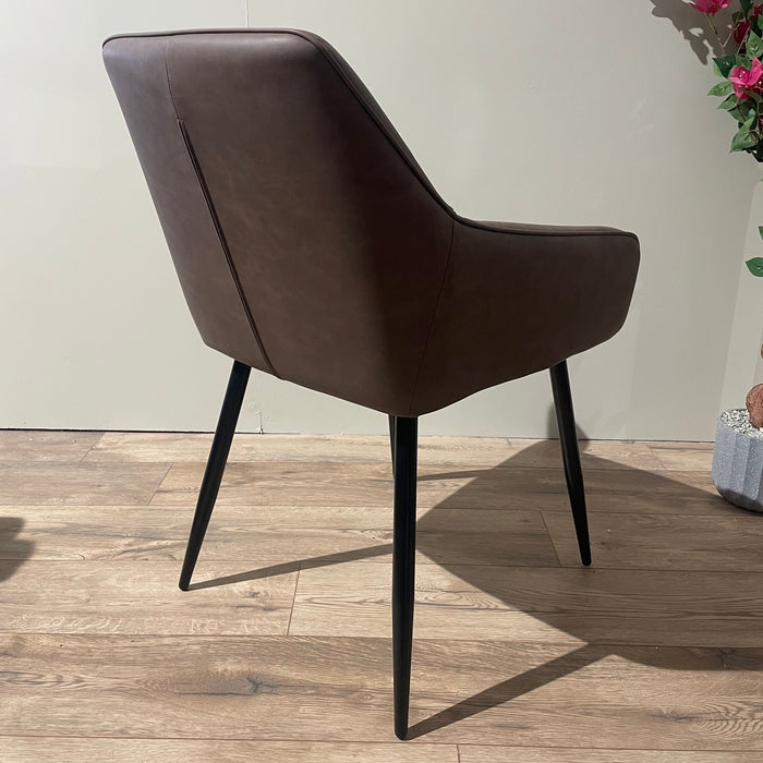 The Enzo Dining Chair features brown leatherette cushioned seats and black metal legs, placed on a wooden floor beside a light wall, with a decorative plant and pink flowers to the right.