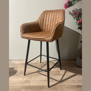 The Enzo Counter Stool, featuring a brown cushioned seat with vertical stitching, black metal legs, and a footrest, stands on a wooden floor beside a potted plant with pink flowers against a light wall.
