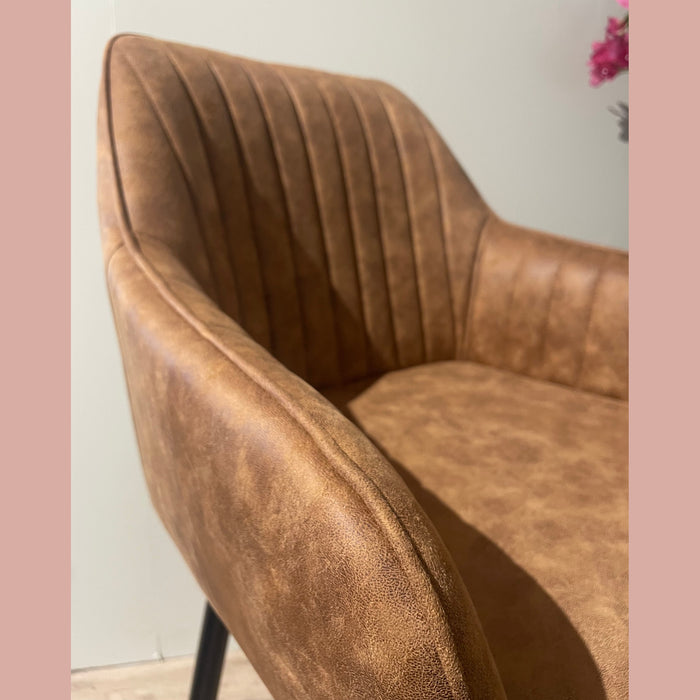 A close-up of the Enzo Counter Stool, a brown faux leather chair with vertical stitching on the backrest and armrests, set against a light wall with pink flowers visible in the top right corner.