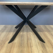 A close-up of the Elson 1.6 Dining Table, a modern piece featuring a wooden top and bold black X legs, set on a light wood floor with a gray wall in the background.