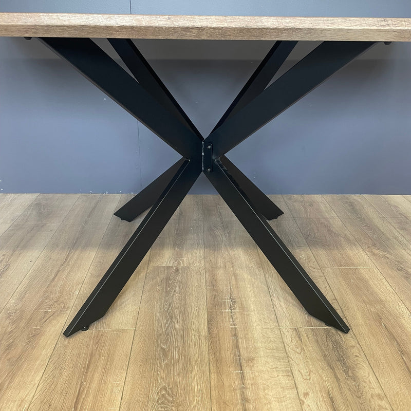 A close-up of the Elson 1.6 Dining Table, a modern piece featuring a wooden top and bold black X legs, set on a light wood floor with a gray wall in the background.