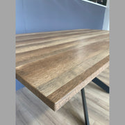 Close-up of the Elson 1.6 Dining Table, a modern piece with a smooth natural wood finish, visible grain, and bold black X legs. Shown on a wooden floor with a light blue wall in the background.