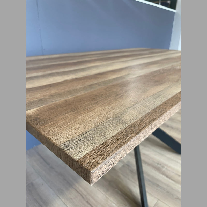 Close-up of the Elson 1.6 Dining Table, a modern piece with a smooth natural wood finish, visible grain, and bold black X legs. Shown on a wooden floor with a light blue wall in the background.