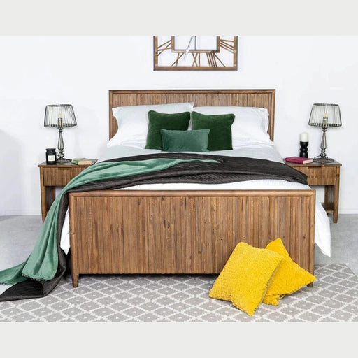 The Cairo Bedframe, made of reclaimed pine, features white bedding, green pillows, and a green and black throw. It’s placed between rustic nightstands, with two yellow pillows on a gray rug and a clock above the headboard.