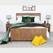 The Cairo Bedframe, made of reclaimed pine, features white bedding, green pillows, and a green and black throw. It’s placed between rustic nightstands, with two yellow pillows on a gray rug and a clock above the headboard.