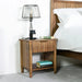 The Cairo Bedside Locker from a rustic collection sits beside the bed, topped with a wired metal lamp, candle jar, and two books. The bed features white bedding and a green pillow, with soft carpeting below.