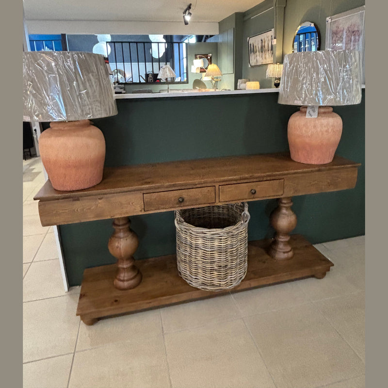 The Leeds Console Table, featuring two central drawers and a wicker basket on the lower shelf, is styled in a modern, well-lit space with two large terracotta lamps wrapped in plastic on either end, radiating rustic charm.