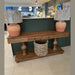 The Leeds Console Table, featuring two central drawers and a wicker basket on the lower shelf, is styled in a modern, well-lit space with two large terracotta lamps wrapped in plastic on either end, radiating rustic charm.