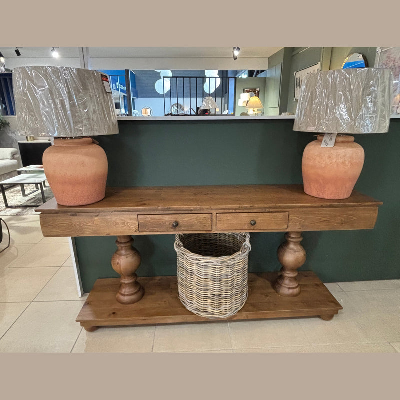 The Leeds Console Table, crafted from reclaimed pine with two drawers and turned legs, brings rustic charm. It’s topped with two large terracotta lamps with wrapped shades, while a wicker basket rests on the lower shelf.