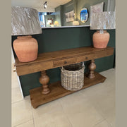 The Leeds Console Table with two drawers displays two large terracotta lamps with wrapped shades. A wicker basket rests on its lower shelf. The table stands against a green wall in a showroom filled with mirrors and framed art.