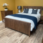 A cozy bedroom with the Cairo Bedframe, white bedding, dark gray pillows, blue throw, rustic nightstands and dresser, a silver lamp, and a potted orchid set against a yellow wall.