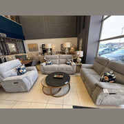 A furniture showroom features three gray Barrington Electric Recliners with supportive cushions, arranged around round coffee tables. Floral pillows, lamps, wall art, and decor enhance the modern space filled with natural light from large windows.