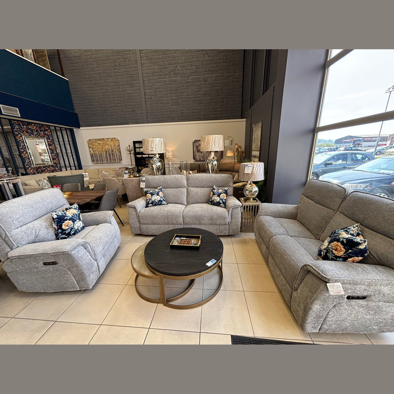 A furniture showroom features three gray Barrington Electric Recliners with supportive cushions, arranged around round coffee tables. Floral pillows, lamps, wall art, and decor enhance the modern space filled with natural light from large windows.