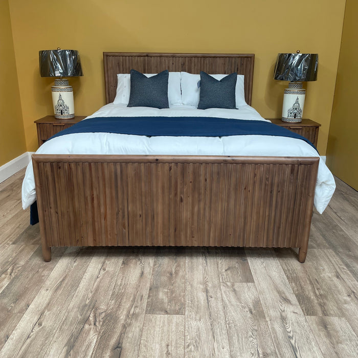 The Cairo Bedframe, made from reclaimed pine, features white bedding and dark blue pillows against a yellow wall. Two matching nightstands with black lamps and decorative bases complete this rustic bedroom furniture set.